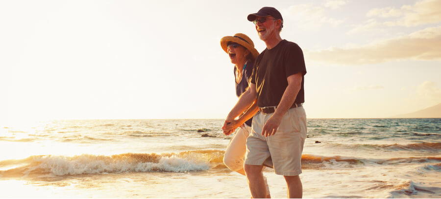 Un couple âgé riant et marchant ensemble sur une plage au bord de l'eau.